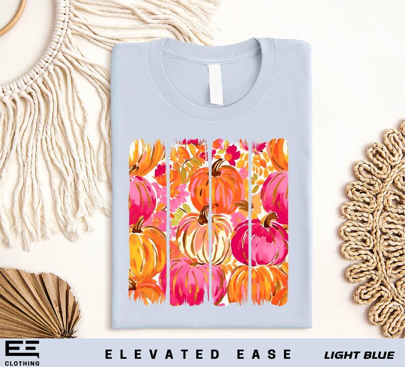 a light blue t-shirt with a vibrant graphic design of orange and pink pumpkins, arranged in a grid pattern. The design is displayed on a white background, and there are additional decorative elements such as a woven basket and a macrame wall hanging in the background.