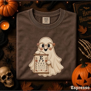 May include: A brown crew neck t-shirt featuring an embroidered graphic of a ghost wearing floral glasses and holding an eye chart with the letters E, F, P, T, O, Z, L, P, E, D, P, E, C, F, D, K, D, F, C, Z. The shirt has a Comfort Colors brand label.