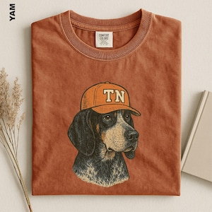 May include: A rust-colored t-shirt featuring a graphic illustration of a hound dog wearing an orange baseball cap with the letters TN printed on the front. The shirt has a crew neckline and a Comfort Colors brand label inside the collar.