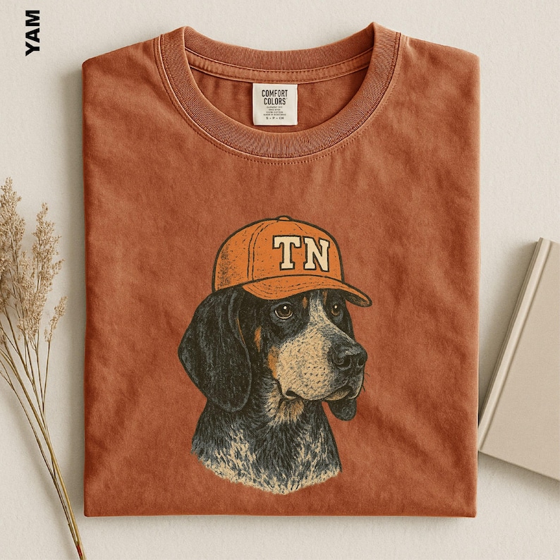 May include: A rust-colored t-shirt featuring a graphic illustration of a hound dog wearing an orange baseball cap with the letters TN printed on the front. The shirt has a crew neckline and a Comfort Colors brand label inside the collar.
