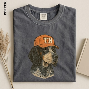 May include: A charcoal gray crew neck t-shirt featuring a graphic illustration of a hound dog wearing an orange baseball cap with the letters TN printed on the front. The shirt has a visible brand label on the inside collar area.