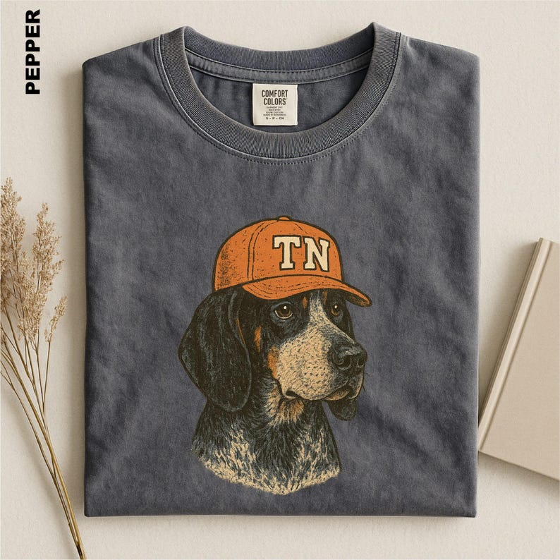 May include: A charcoal gray crew neck t-shirt featuring a graphic illustration of a hound dog wearing an orange baseball cap with the letters TN printed on the front. The shirt has a visible brand label on the inside collar area.