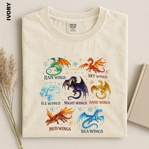 May include: An ivory t-shirt featuring a graphic print of seven colorful dragons arranged in a circle. Each dragon is labeled with text including Rain Wings, Sky Wings, Ice Wings, Night Wings, Sand Wings, Mud Wings, and Sea Wings.