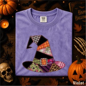 May include: A purple t-shirt featuring a central graphic of a witch hat constructed from various fabric patches, including plaid, leopard print, and floral patterns. The hat is outlined with a decorative stitch and includes a central buckle detail.