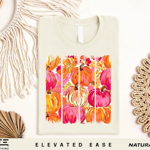 a white t-shirt with a vibrant graphic design of pumpkins in shades of orange, pink, and yellow. The pumpkins are arranged in a square pattern, creating a visually appealing and festive design. The t-shirt is displayed on a white background, and there are additional pumpkins and a woven basket visible in the background. The phrase "Elevated Ease" and "Natural" are also present in the image.