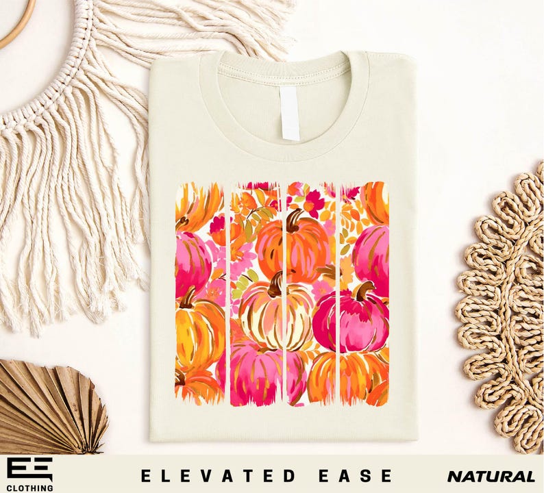 a white t-shirt with a vibrant graphic design of pumpkins in shades of orange, pink, and yellow. The pumpkins are arranged in a square pattern, creating a visually appealing and festive design. The t-shirt is displayed on a white background, and there are additional pumpkins and a woven basket visible in the background. The phrase "Elevated Ease" and "Natural" are also present in the image.