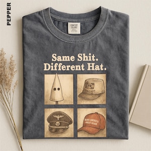 May include: A charcoal gray crew neck t-shirt featuring the text "Same Shit. Different Hat." above four square illustrations of various headwear, including a pointed hood, a military-style cap, a peaked cap with an emblem, and a red baseball cap.