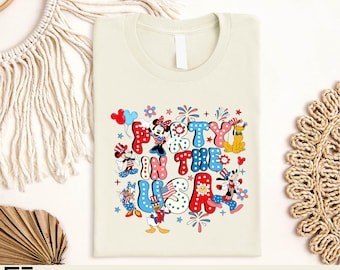 Può includere: T-shirt color crema con la scritta "Party in the USA" in rosso, bianco e blu, con personaggi dei cartoni animati. Il design include Topolino e Minnie, Paperino e Pluto, con stelle e fuochi d'artificio.