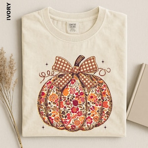 May include: An ivory t-shirt featuring a central graphic of a pumpkin filled with a floral pattern in shades of orange, red, and brown. The pumpkin is topped with a brown and white gingham bow and surrounded by small four-pointed star accents.