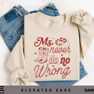 May include: A sand-colored crewneck sweatshirt featuring a red graphic print of a cowboy hat, a pair of cowboy boots, and the text "Ms. never do no Wrong" in a stylized font, layered over a pair of blue denim jeans.
