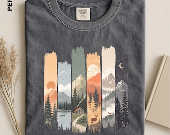 May include: A charcoal gray t-shirt featuring a graphic print of five vertical panels depicting a mountain landscape transition from sunrise to night. The design includes pine trees, a lake with a boat, a cabin, a deer, and a crescent moon.