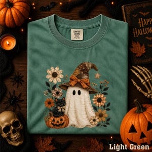May include: A sage green t-shirt featuring a graphic of a white ghost wearing a brown plaid witch hat, sitting next to a black cat on a pumpkin, surrounded by various orange and brown flowers, with a Comfort Colors brand tag at the neck.
