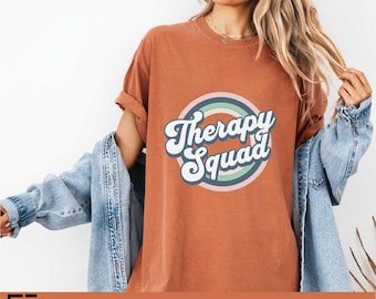 May include: A rust-colored short-sleeve t-shirt featuring a graphic design with the text Therapy Squad in white cursive lettering, centered within a series of concentric circles in muted shades of pink, green, and blue, worn with a denim jacket.