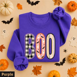 May include: A purple long-sleeve sweatshirt featuring the word BOO printed in large letters. The B is black and white buffalo plaid, the O is pink with small ghosts and bats, and the second O has vertical orange, purple, and white stripes.