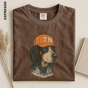 May include: A dark brown crew neck t-shirt featuring a graphic illustration of a hound dog wearing an orange baseball cap with the letters TN printed on the front. The shirt has a Comfort Colors brand label inside the collar and a vintage wash.
