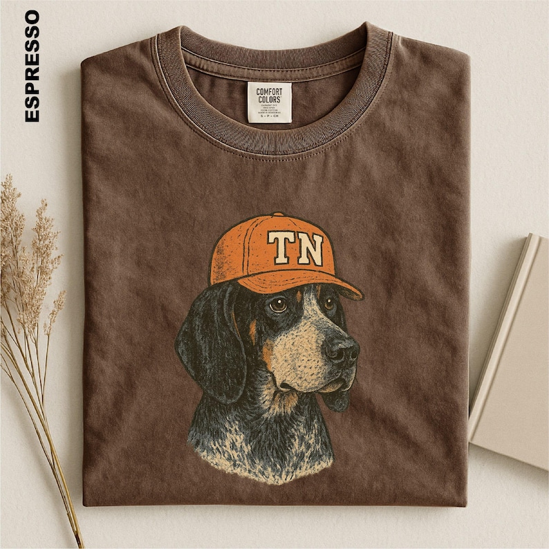 May include: A dark brown crew neck t-shirt featuring a graphic illustration of a hound dog wearing an orange baseball cap with the letters TN printed on the front. The shirt has a Comfort Colors brand label inside the collar and a vintage wash.