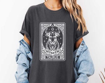 Puede incluir: Una camiseta gris oscuro con un gráfico blanco de un signo zodiacal de Tauro. El gráfico presenta un toro con un diseño de estrella y luna creciente. El texto "Tauro" está debajo del toro.