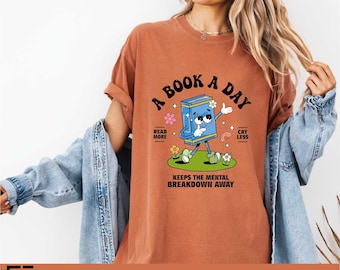 May include: A rust-colored t-shirt featuring a graphic of a blue cartoon book character walking on green grass. The text reads A BOOK A DAY, READ MORE, CRY LESS, and KEEPS THE MENTAL BREAKDOWN AWAY, surrounded by small flowers, stars, and a rainbow.