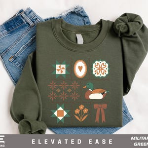 May include: Military green sweatshirt with a decorative design featuring a duck, flowers, and geometric shapes. The sweatshirt is paired with blue jeans. The text "ELEVATED EASE" and "MILITARY GREEN" are visible.