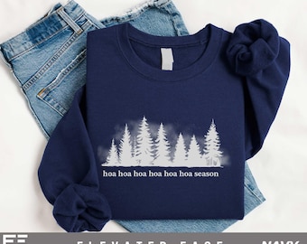 May include: A navy blue long-sleeve sweatshirt featuring a white graphic of a forest of pine trees with a misty effect. Below the trees, the text "hoa hoa hoa hoa hoa hoa season" is printed in white. The sweatshirt is folded over blue denim jeans.