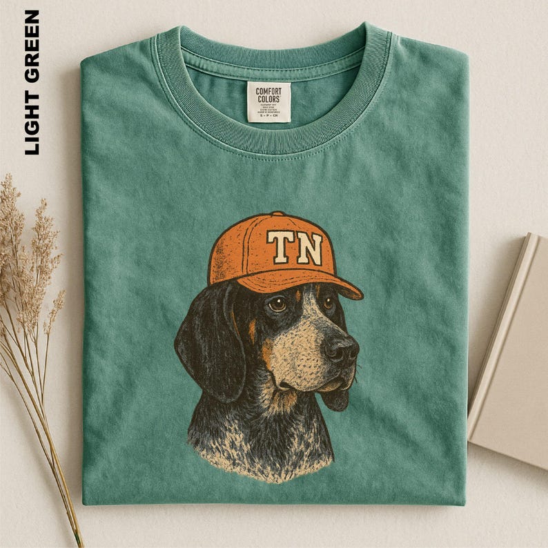 May include: A light green crew neck t-shirt featuring a graphic illustration of a dog wearing an orange baseball cap with the letters TN printed on the front. The shirt has a Comfort Colors brand label on the inside of the collar.