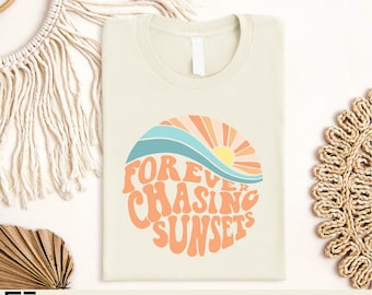 May include: Cream-colored t-shirt with a retro graphic of a sunset over ocean waves. The design features the text "FOREVER CHASING SUNSETS" in orange, with blue, orange, and yellow accents. The shirt is made of a soft material.