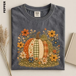 May include: A charcoal gray t-shirt featuring a central graphic of a pumpkin composed of various patterned fabric segments in orange, green, and cream. The pumpkin is surrounded by embroidered-style flowers in yellow, orange, and blue.