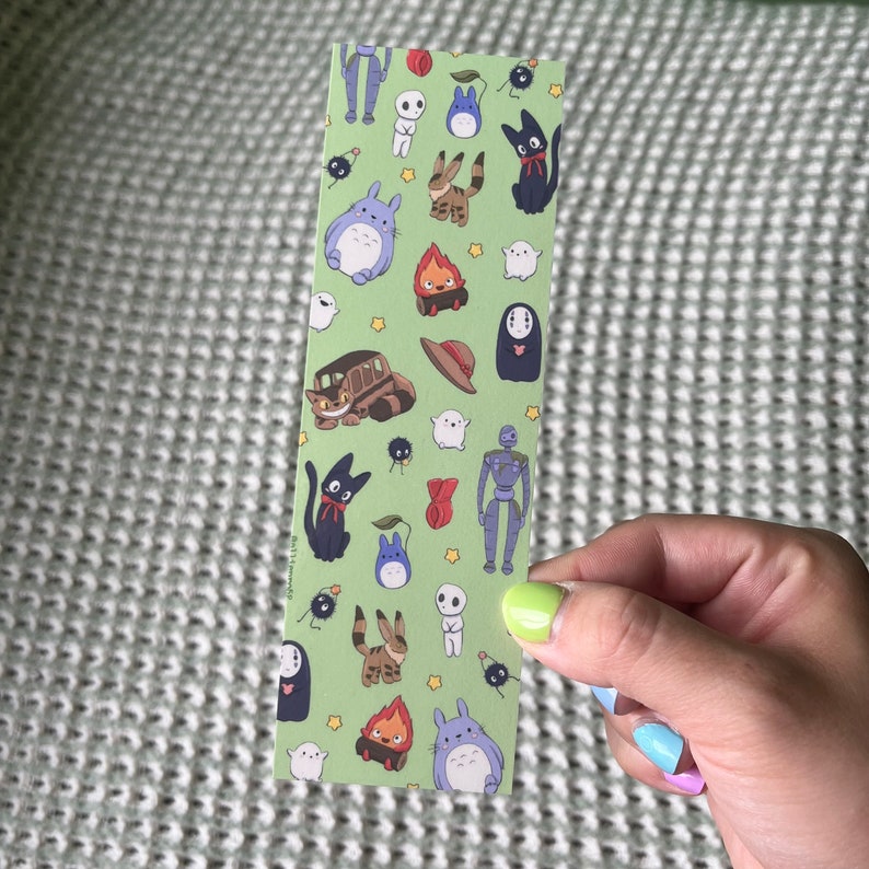 May include: A green bookmark featuring various cartoon characters in a repeating pattern. The characters include a large blue creature, a black cat, a train, and other whimsical figures. The bookmark is held by a person.