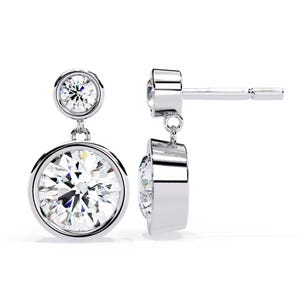 May include: A pair of elegant, silver-toned drop earrings. Each earring features a large, round, clear gemstone set in a bezel setting, with a smaller gemstone above. The earrings are designed with a post and dangle style, showcasing the sparkling stones.