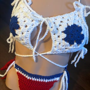 May include: A handmade crochet bikini set in red, white, and blue. The top is white with blue floral accents and tie closures. The bottoms are red with blue and white trim and tie sides. This swimwear is perfect for summer.