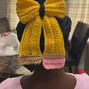 May include: A handmade, yellow crochet hair bow in the shape of a pencil. The bow is attached to a black hair tie. The pencil design features a brown eraser, a gold band, and a pink tip. The bow is worn in the hair.