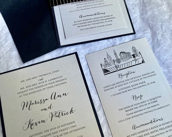 Puede incluir: Un conjunto de invitaciones de boda con detalles en negro y dorado. La invitación presenta una ilustración de la ciudad y el texto "Marissa Ann y Kevin Patrick". La tarjeta de recepción presenta una ilustración de la ciudad y el texto "Recepción".