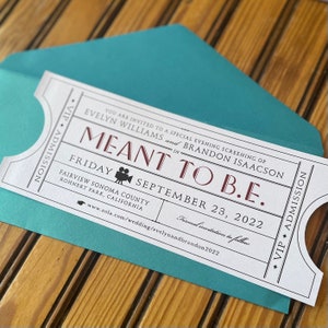 May include: A white invitation card with a movie ticket design, featuring the text "MEANT TO B.E." and event details. The card is inside a teal envelope, set against a wooden surface.