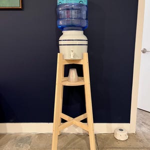 May include: A wooden water cooler stand with a white ceramic water dispenser and a blue plastic water bottle. The bottle has a blue and turquoise label that reads "Primo Water".