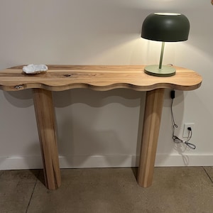 May include: A wooden console table with a wavy top and two cylindrical legs. A small white dish sits on the table, and a green lamp with a round shade is on the right side of the table.