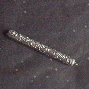 Può includere: Un ciondolo cilindrico color argento con intricati motivi floreali e a spirale. Il ciondolo ha un piccolo anello a un'estremità. Lo sfondo è scuro con piccoli punti bianchi e iridescenti sparsi.