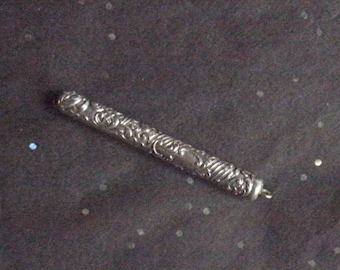 May include: A silver-colored, cylindrical pendant with intricate floral and swirl patterns. The pendant has a small loop at one end. The background is dark with small, scattered, white and iridescent specks.