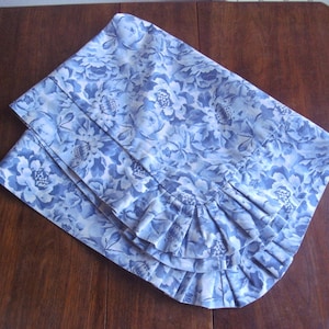 Può includere: Un tessuto floreale blu e bianco con un bordo arricciato. Il tessuto è piegato a metà e ha uno sfondo blu chiaro con fiori bianchi.