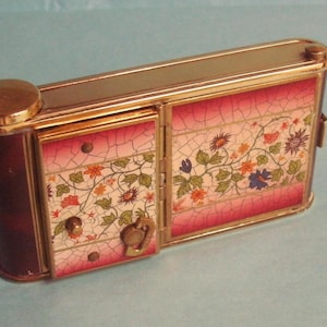 Könnte beinhalten: Ein rechteckiger Vintage-Kompakt mit floralem Design. Der Kompakt hat eine burgunderfarbene zylindrische Seite und einen goldfarbenen Rahmen. Die Frontplatten zeigen ein rosa-weißes Rissmuster mit farbenfrohen floralen Akzenten.