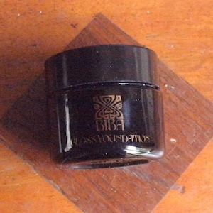 Puede incluir: Un frasco negro de Biba Gloss Foundations está sobre una superficie de madera. El frasco tiene un emblema y texto dorados. El frasco es cilíndrico con una tapa plana y negra. El fondo es una superficie de madera con una incrustación en forma de diamante.