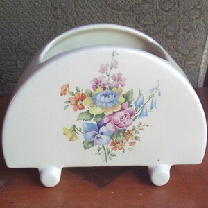 Puede incluir: Un macetero de cerámica blanca con un diseño floral. El macetero tiene forma de media luna y tres pequeños pies. Las flores están pintadas en tonos de rosa, azul, amarillo y naranja.