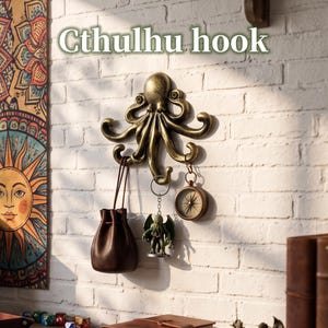 Op de afbeelding: Een aan de muur gemonteerde haak in de vorm van een octopus met een metallic gouden afwerking. De haak heeft meerdere gebogen tentakels voor het ophangen van voorwerpen. Een bruin lederen tasje, een Cthulhu-sleutelhanger en een rond messing kompas hangen eraan.