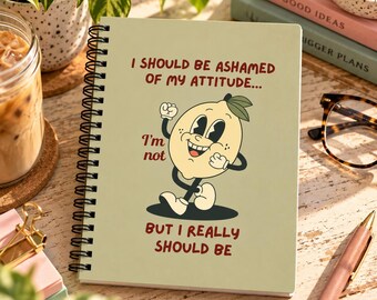 May include: A spiral-bound notebook with a light green cover featuring a cartoon lemon character and the text: I SHOULD BE ASHAMED OF MY ATTITUDE... I'm not BUT I REALLY SHOULD BE. The lemon has a smiling face, arms, and legs.