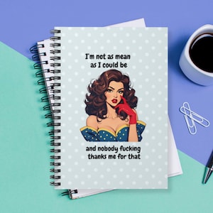 May include: A spiral-bound notebook with a light blue cover featuring white polka dots. The cover displays a retro illustration of a woman with text that reads, "I'm not as mean as I could be and nobody fucking thanks me for that."