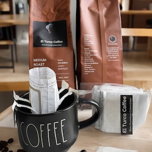 May include: A black coffee mug with the word "COFFEE" written on it. The mug is filled with single-serve coffee pods. Two bags of El Turco Coffee are in the background, one with the text "El Turco Coffee Pure Premium Colombian" and the other with the text "MEDIUM ROAST ORIGIN COLOMBIA VARIETY CASTILLO MAY, 2024".