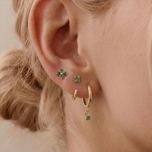 Puede incluir: Primer plano de una oreja adornada con pendientes dorados. La oreja presenta un pequeño y cuadrado pendiente verde, un pequeño aro dorado y un aro dorado más grande con una gema verde colgante. También se ve un pendiente en forma de trébol de cuatro hojas con gemas verdes.
