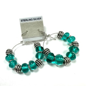 A pair of sterling silver drop hoop earrings featuring handmade turquoise green glass beads made using a torch and kiln. The total length is approximately 2 inches.