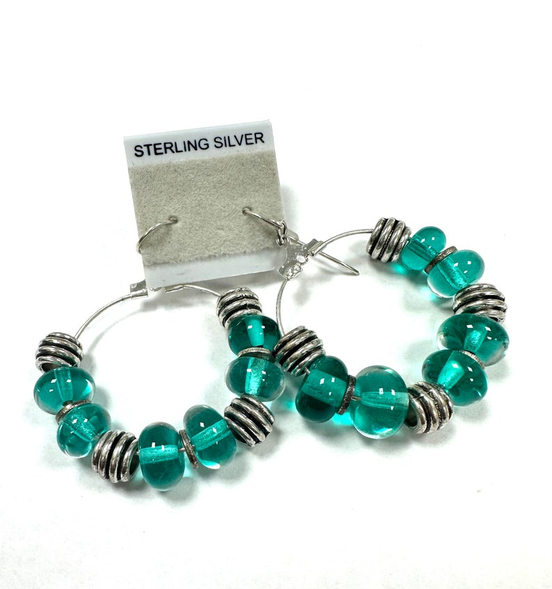 A pair of sterling silver drop hoop earrings featuring handmade turquoise green glass beads made using a torch and kiln. The total length is approximately 2 inches.