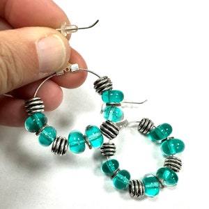 Holding one of the sterling silver hoops featuring handmade transparent turquoise green glass beaded earrings.