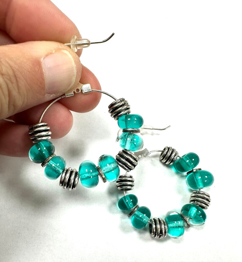 Holding one of the sterling silver hoops featuring handmade transparent turquoise green glass beaded earrings.